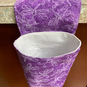 May include: A purple fabric hanging trash can with a matching lid. The can and lid are decorated with a white dragonfly pattern. The interior of the can is white with a woven texture. The can is hanging on a dark brown surface.