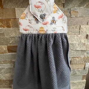 May include: A hanging kitchen towel featuring a dark gray textured fabric base attached to a white fabric top with a bee, honeycomb, and floral print. The top section is secured with a gray snap button and hangs from a wooden rod.