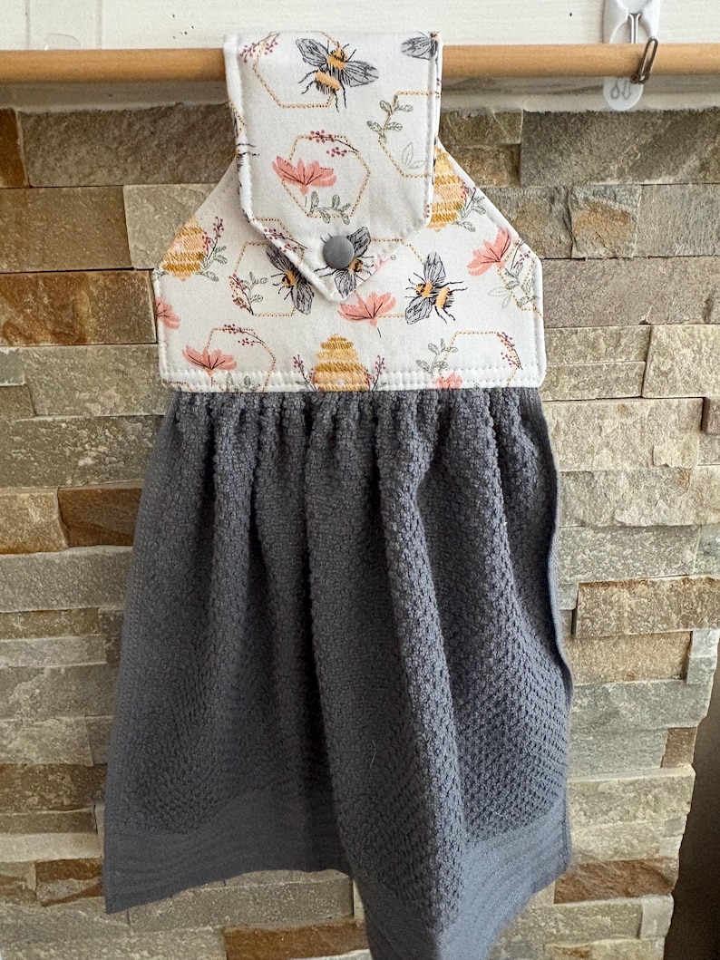 May include: A hanging kitchen towel featuring a dark gray textured fabric base attached to a white fabric top with a bee, honeycomb, and floral print. The top section is secured with a gray snap button and hangs from a wooden rod.