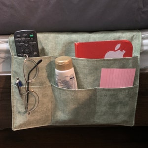 May include: A green fabric bed caddy with multiple pockets. The caddy is hanging on the side of a bed and contains a remote control, glasses, a bottle of lotion, a notebook, and a pink notepad.