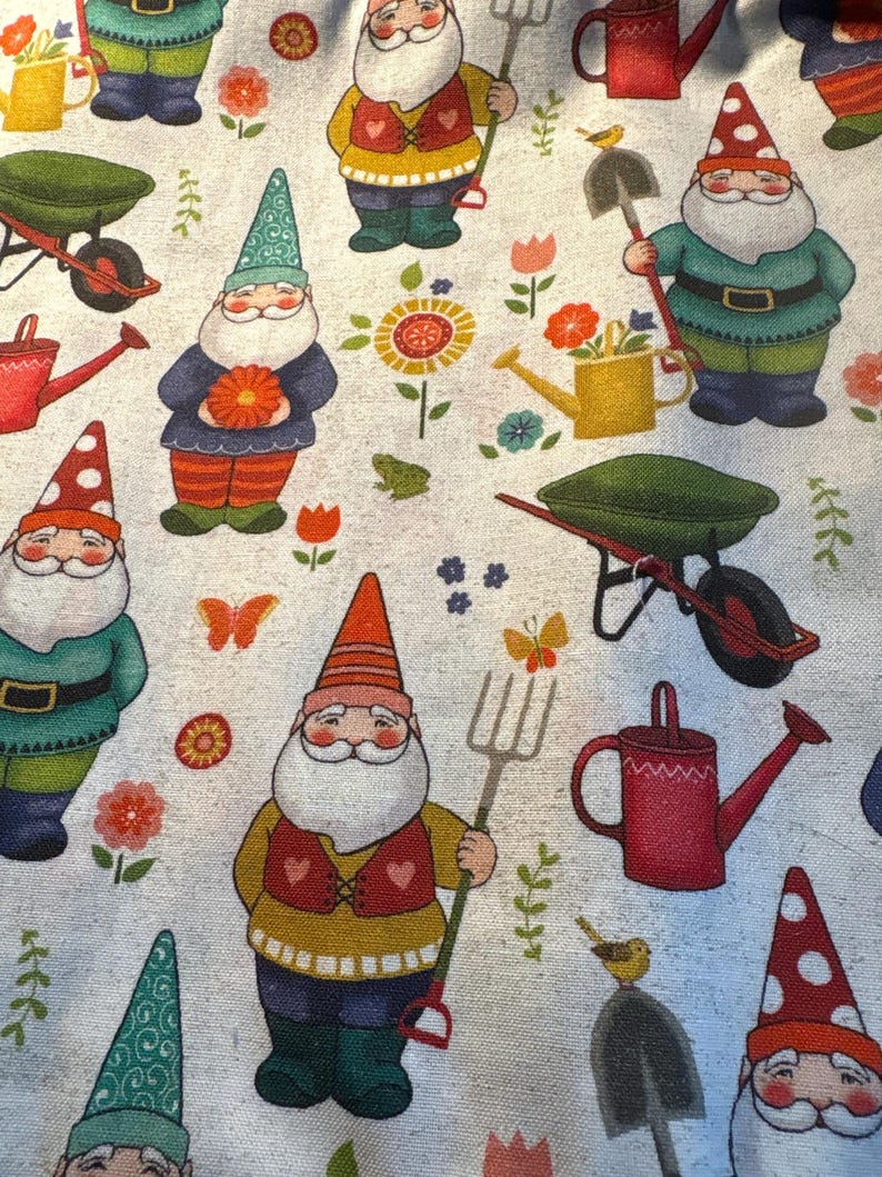 May include: A light gray fabric featuring a repeating pattern of garden gnomes with white beards, colorful hats, and vests. The design includes gardening tools like shovels, pitchforks, red watering cans, green wheelbarrows, flowers, and butterflies.