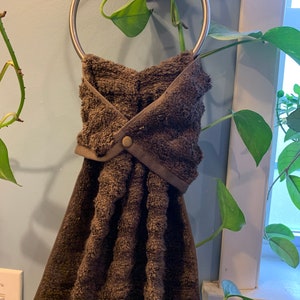 May include: A brown kitchen towel is hanging on a silver towel ring. The towel is folded in a way that creates a pocket for the towel to hang on the ring. The towel has a button on the front.