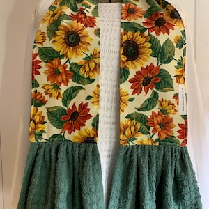 May include: A green tea towel with a sunflower print hanging on a white background. The towel has a ruffle at the bottom and a loop at the top.