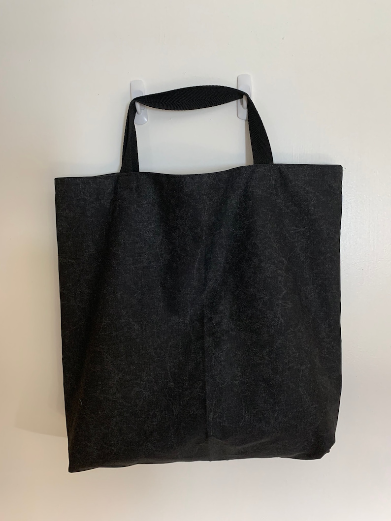 May include: A black canvas tote bag with black fabric handles. The bag is hanging on a white wall.