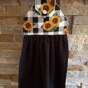 May include: A hanging kitchen towel featuring a black solid-colored textured fabric attached to a fabric topper. The topper displays a black and white buffalo check pattern decorated with yellow sunflower prints and a single white plastic snap button.
