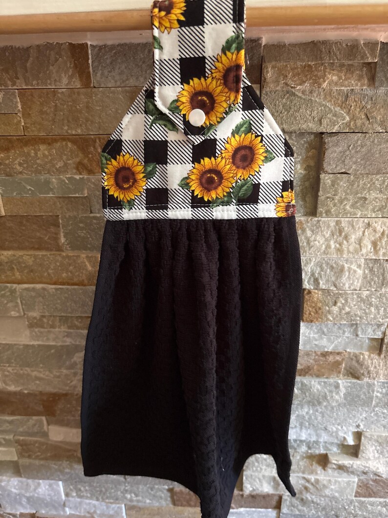 May include: A hanging kitchen towel featuring a black solid-colored textured fabric attached to a fabric topper. The topper displays a black and white buffalo check pattern decorated with yellow sunflower prints and a single white plastic snap button.