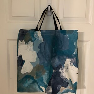 May include: A blue and white tote bag with an abstract floral pattern. The bag has black handles and is hanging on a white hook.