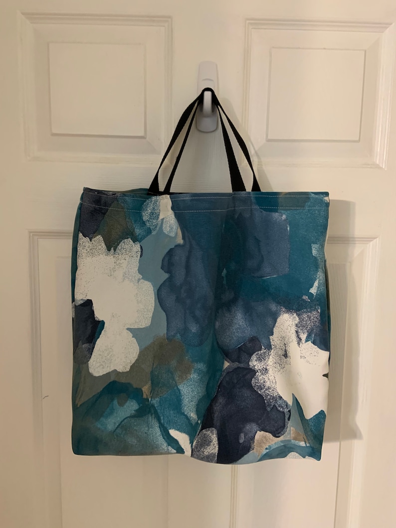 May include: A blue and white tote bag with an abstract floral pattern. The bag has black handles and is hanging on a white hook.