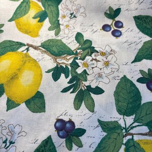 May include: A white fabric featuring a repeating pattern of bright yellow lemons, clusters of small dark purple berries, and white blossoms with green leaves. The background is overlaid with faint, cursive black script text throughout the design.