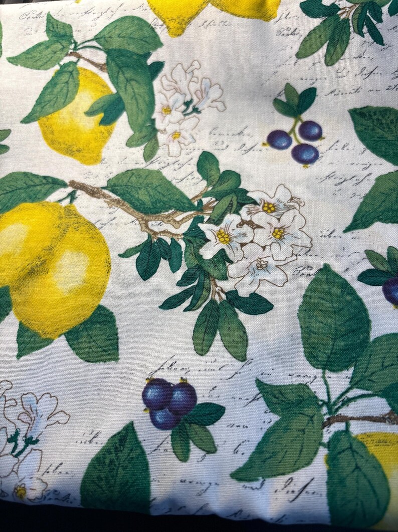 May include: A white fabric featuring a repeating pattern of bright yellow lemons, clusters of small dark purple berries, and white blossoms with green leaves. The background is overlaid with faint, cursive black script text throughout the design.