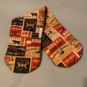 May include: A pair of red and black oven mitts with a BBQ theme. The mitts are covered in vintage-style advertisements for barbecue, grilling, and smoking meat. The text on the mitts includes phrases like "Backyard BBQ Master", "The King of BBQ", and "Almost Heaven!"