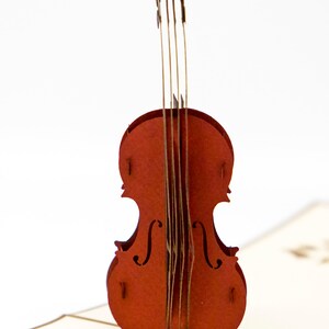 May include: A red papercraft pop-up card featuring a detailed violin. The violin is made of layered paper and has a realistic design.