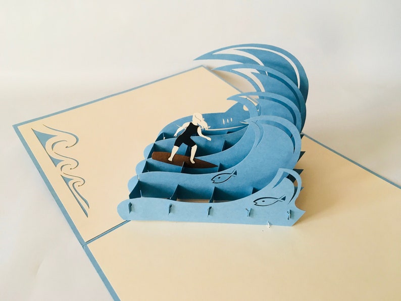 May include: A blue and white 3D pop-up card featuring a surfer riding a wave with two fish in the background.