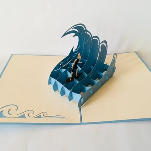 May include: A blue 3D pop-up card featuring a person surfing on a wave. The card is open and the wave is raised up. The card is on a cream colored background.