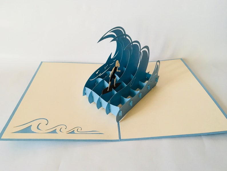 May include: A blue 3D pop-up card featuring a person surfing on a wave. The card is open and the wave is raised up. The card is on a cream colored background.