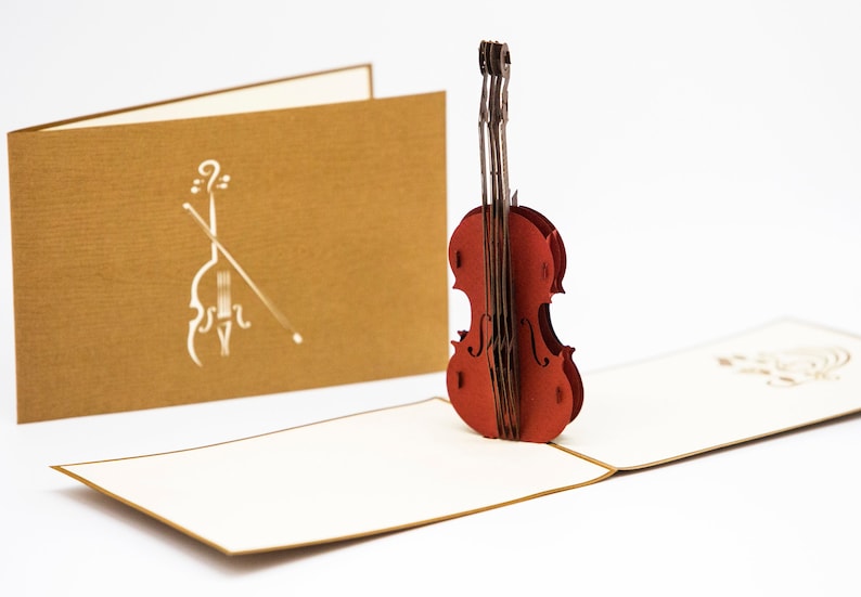 May include: A red pop-up card featuring a 3D violin. The card is open and the violin is standing up. The card has a brown outer layer and a white inner layer.
