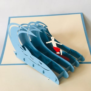 May include: A blue 3D pop-up card featuring a surfer riding a wave. The card is made of paper and has a detailed design.