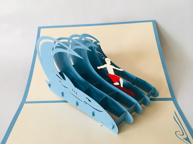 May include: A blue 3D pop-up card featuring a surfer riding a wave. The card is made of paper and has a detailed design.