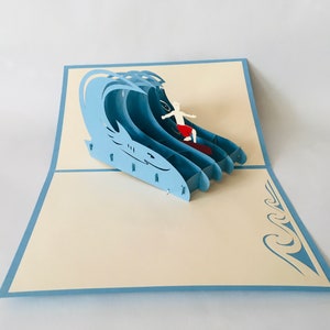 May include: A blue 3D pop-up card featuring a surfer riding a wave. The card is open and the surfer is visible in the center of the card. The card is on a white background.