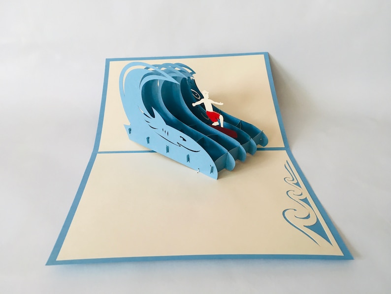 May include: A blue 3D pop-up card featuring a surfer riding a wave. The card is open and the surfer is visible in the center of the card. The card is on a white background.