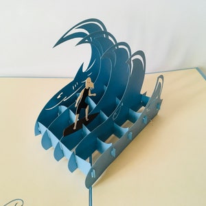 May include: A blue paper pop-up card featuring a person surfing on a wave. The card is made of paper and has a 3D effect.