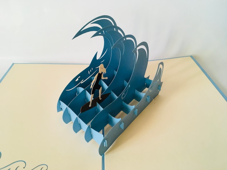 May include: A blue paper pop-up card featuring a person surfing on a wave. The card is made of paper and has a 3D effect.