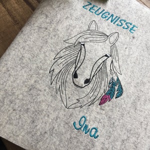 May include: Gray felt folder with a black and white embroidered horse head with pink and blue feathers. The text "ZEUGNISSE" is embroidered in blue above the horse and the name "Ina" is embroidered in blue below the horse.