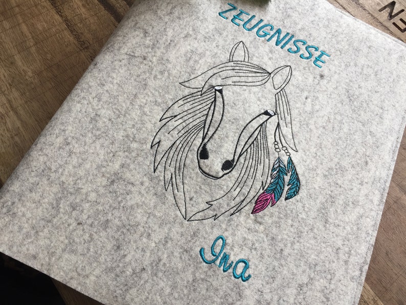 May include: Gray felt folder with a black and white embroidered horse head with pink and blue feathers. The text "ZEUGNISSE" is embroidered in blue above the horse and the name "Ina" is embroidered in blue below the horse.