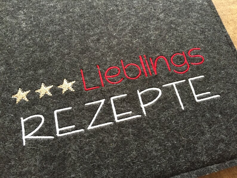 May include: Dark gray felt recipe holder with embroidered text. The word "Lieblings" is in red, and "Rezepte" is in white. Three gold stars are embroidered above the word "Lieblings".