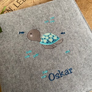 May include: A gray felt baby blanket with a blue and green embroidered turtle and the name "Oskar" in blue embroidery.