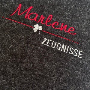Könnte beinhalten: Ein schwarzer Filzordner mit dem Namen "Marlene" in rot und dem Wort "ZEUGNISSE" in weiß gestickt.