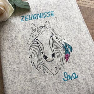 May include: Gray felt folder with a black and white embroidered horse head design. The horse has turquoise and pink feathers and beads. The text "ZEUGNISSE" is embroidered in turquoise above the horse and the name "Ina" is embroidered in turquoise below the horse.