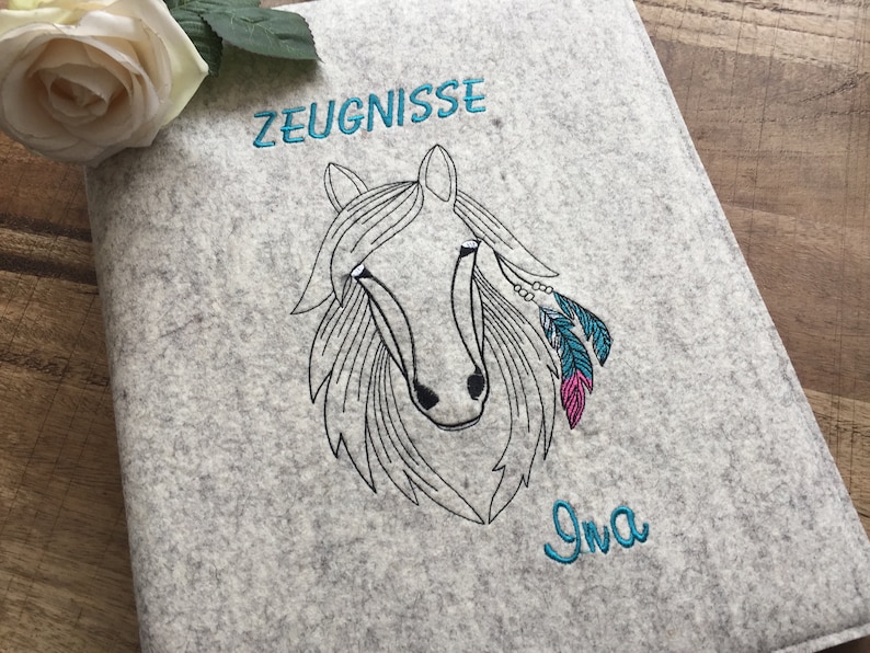 May include: Gray felt folder with a black and white embroidered horse head design. The horse has turquoise and pink feathers and beads. The text "ZEUGNISSE" is embroidered in turquoise above the horse and the name "Ina" is embroidered in turquoise below the horse.