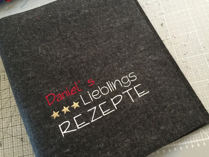 May include: Dark gray felt recipe book cover with embroidered text. The text reads "Daniel's Lieblings REZEPTE" in white and red thread, with three gold stars. A handmade item.