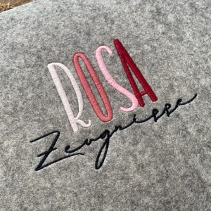 May include: A gray felt fabric with the name "ROSA" embroidered in pink, red, and white lettering. The word "Zweignere" is embroidered in black cursive lettering below the name.