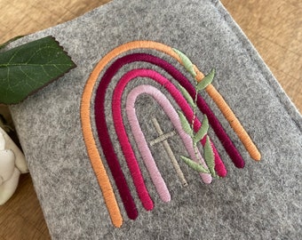 May include: A gray felt book cover with an embroidered rainbow design. The rainbow is made up of orange, red, pink, and purple stripes. A silver cross is embroidered in the center of the rainbow, and green leaves are embroidered on the right side of the rainbow.