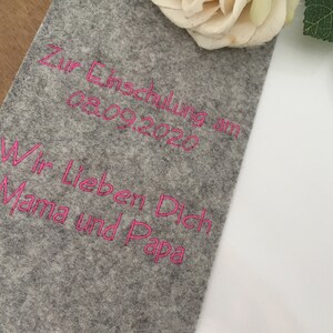 May include: A gray felt fabric with pink embroidered text that reads "Zur Einschulung am 08.09.2020 Wir Lieben Dich Mama und Papa".