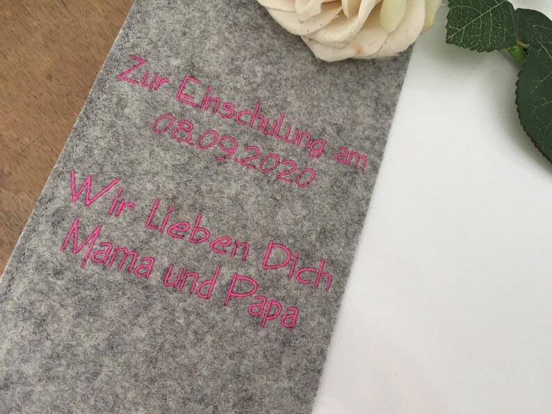 May include: A gray felt fabric with pink embroidered text that reads "Zur Einschulung am 08.09.2020 Wir Lieben Dich Mama und Papa".