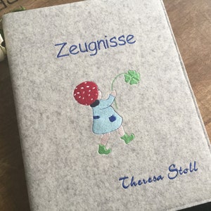 Könnte beinhalten: Ein grauer Filzordner mit dem Wort "Zeugnisse" in blau gestickt. Ein Mädchen mit einem roten Pilzhut und einem blauen Kleid hält einen vierblättrigen Klee. Der Name "Theresa Stoll" ist unten in blau gestickt.