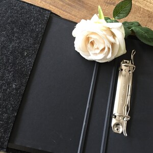 May include: A black binder with a gray felt cover, open to reveal a metal clasp. A white rose with green leaves rests on the binder. The binder is on a wooden surface.