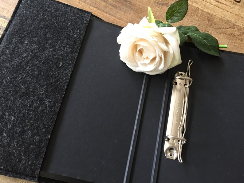 May include: A black binder with a gray felt cover, open to reveal a metal clasp. A white rose with green leaves rests on the binder. The binder is on a wooden surface.