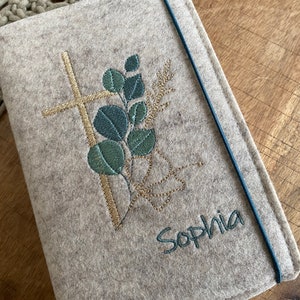 May include: A beige felt book cover with a gold cross and green leaves embroidered on it. The name "Sophia" is embroidered in green below the design.