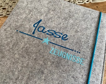 Könnte beinhalten: Grauer Filzordner mit einem blauen Schnallverschluss. Der Ordner trägt den Namen "Lasse" in blauer Stickerei mit einem blauen Stern und dem Wort "Zeugnisse" in blauer Stickerei.