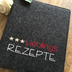May include: A dark gray felt recipe book cover with embroidered text and star accents. The text reads "Lieblings REZEPTE" in white and red thread. A white flower is visible in the background.