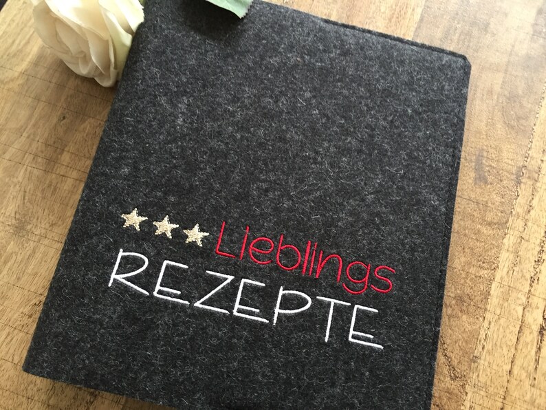 May include: A dark gray felt recipe book cover with embroidered text and star accents. The text reads "Lieblings REZEPTE" in white and red thread. A white flower is visible in the background.