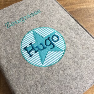 Könnte beinhalten: Grauer Filzordner mit dem Wort "Zeugnisse" obenauf gestickt und einem blauen Kreis mit einem weiß-blau gestreiften Hintergrund und einem blauen Stern mit dem Namen "Hugo" im Kreis gestickt.