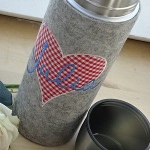 Könnte beinhalten: Eine graue Filz-Isolierflasche mit einem rot-weiß karierten Herzen und dem Wort "Julia" bestickt. Die Flasche hat einen Edelstahldeckel.