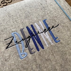 Könnte beinhalten: Ein graues Filz-Notizbuch mit dem Wort "Zenspire" in Schwarz und dem Wort "BE" in Blau, Grau und Weiß gestickt.