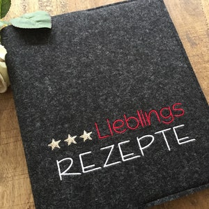 May include: A dark gray felt recipe book cover with the German words "Lieblings REZTE" embroidered in red and white, and three gold stars. A white rose and green leaves are in the upper left corner.