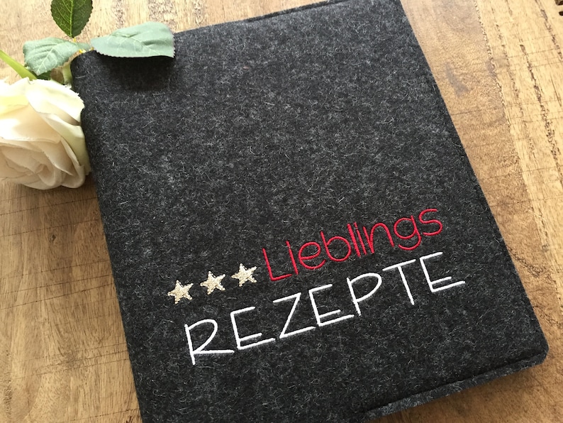 May include: A dark gray felt recipe book cover with the German words "Lieblings REZTE" embroidered in red and white, and three gold stars. A white rose and green leaves are in the upper left corner.